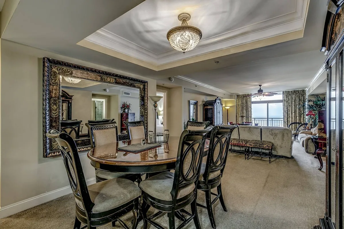 A warm, sophisticated interior shot of the dining area with a chic chandelier, large decorative mirror, and a comfortable sofa that faces the ocean view. The elegant dining table is set for meals, and the space is lit with soft, welcoming light, perfect for celebrations or intimate dinners with family and friends.
