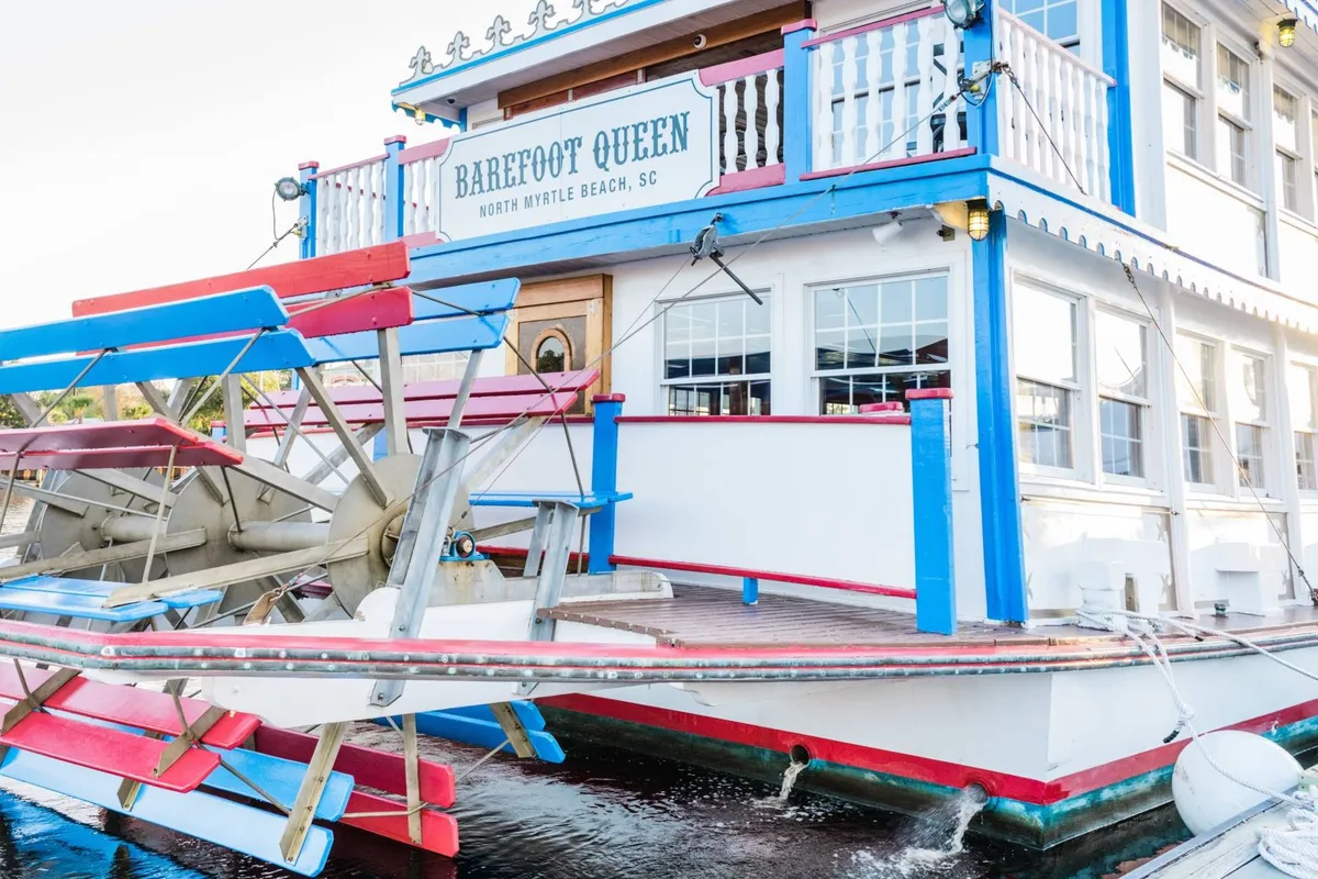 The vibrant exterior of the Barefoot Queen Riverboat is showcased here. Visible are its signature paddle wheel, colorful signs, and festive decor, hinting at the lively sightseeing and on-water dining experiences available nearby. The scene is energetic and inviting, conveying a sense of special adventure.