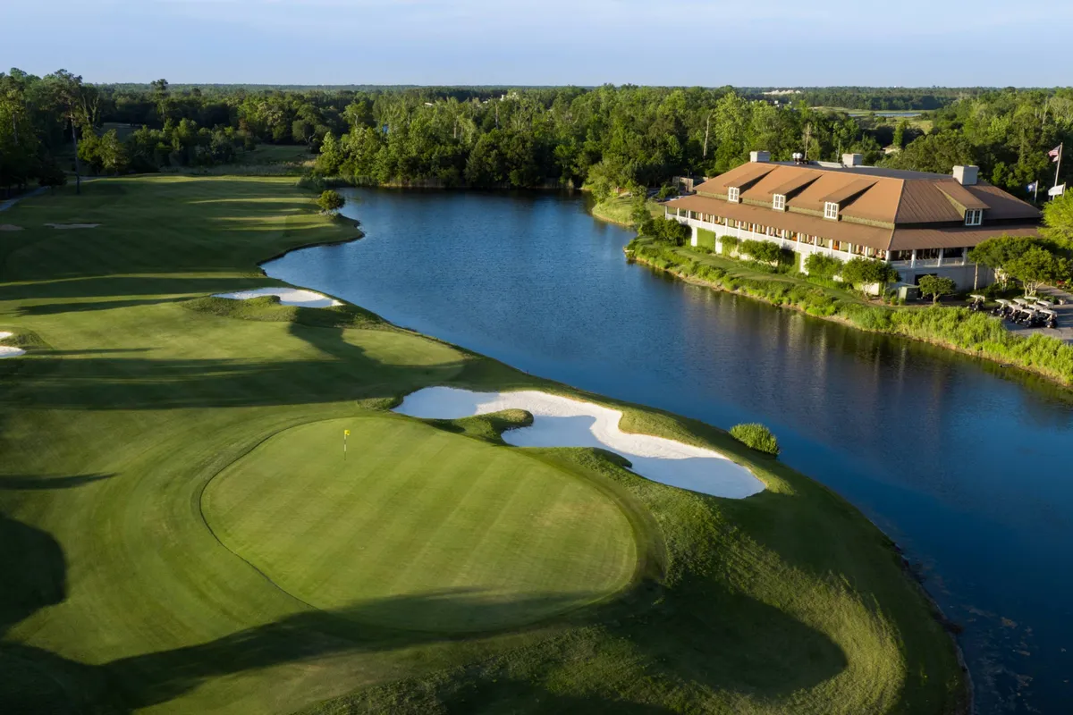 A sprawling golf course bordered by a serene water body and anchored by a prominent clubhouse. Sand traps dot the lush green fairways, while the tranquil surroundings evoke a sense of relaxation and outdoor leisure. The image is bathed in the gentle light of early morning or late afternoon, hinting at both sport and peaceful reprieve.
