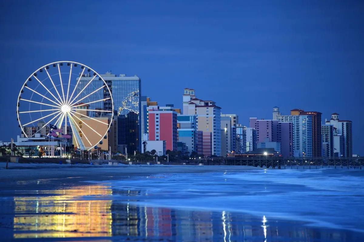 A vibrant nighttime scene features a brightly lit Ferris wheel, colorful beachside buildings, and shimmering ocean reflections. People stroll along the boardwalk, and the overall atmosphere is festive and lively, capturing the spirit of local nightlife and waterfront entertainment.