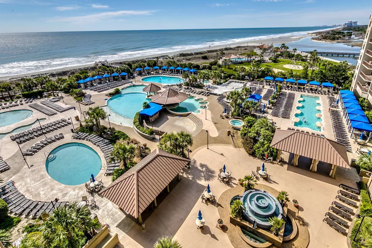 This image features multiple swimming pools framed by palm trees, with lounge chairs scattered around and the beach visible in the background. The scene captures the essence of a vibrant, vacation-like resort atmosphere, highlighting both water features and relaxation zones.