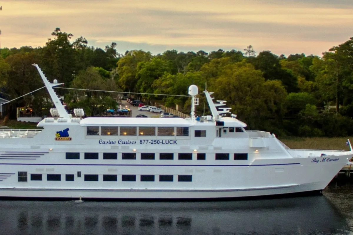 A sunlit exterior shot featuring a large white yacht with clear casino branding docked on a serene waterway, inviting thoughts of relaxation and social experiences. The open deck offers views of the water, the sky, and hints at the excitement within.