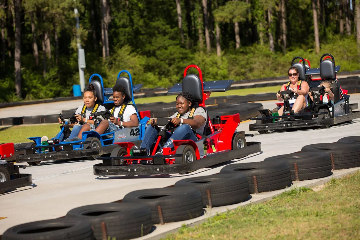 A dynamic exterior scene of the Broadway Grand Prix racetrack, with go-karts in action, participants enjoying the thrill of racing, and lush trees surrounding the track. The energy and excitement are palpable, making it clear this is a destination for family fun and friendly competition.