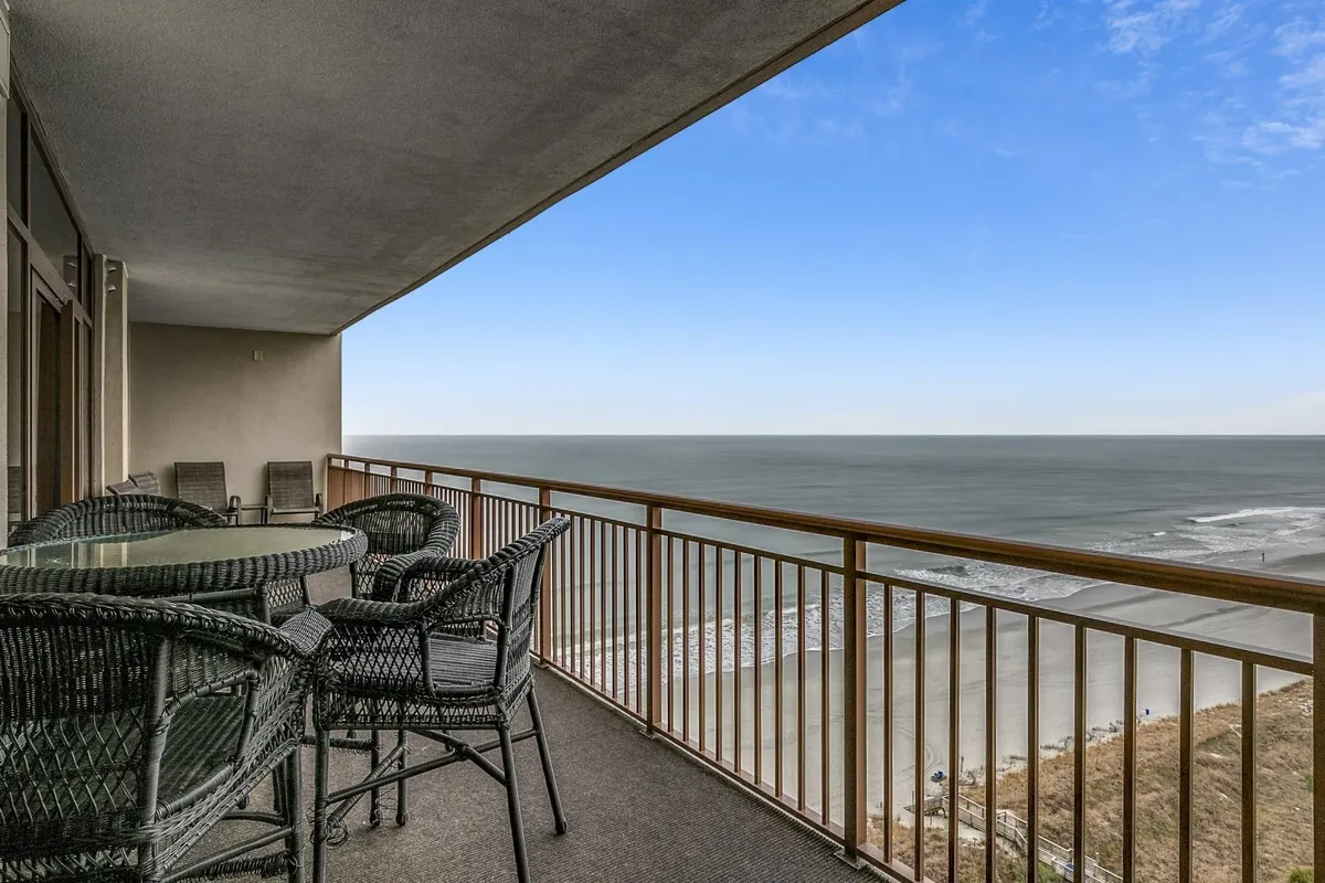 An expansive, covered balcony overlooking the sandy beach and rolling ocean waves. Comfortable outdoor seating is arranged for dining or relaxing while taking in sunrise or sunset vistas. The tranquil mood is set by the proximity to the water and the inviting space for enjoying meals, drinks, or simply the sound of the waves.