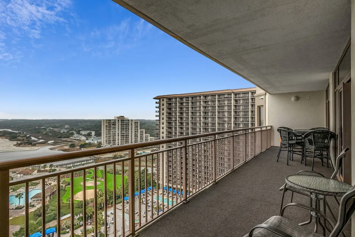 This image captures a serene exterior balcony scene, featuring a sturdy railing, comfortable outdoor seating, and sweeping views of the surrounding landscape—likely providing sightlines to both the beach and pool complex. The peaceful ambiance suggests a perfect spot for families to enjoy sunrise coffee or sunset conversations after a day of fun.