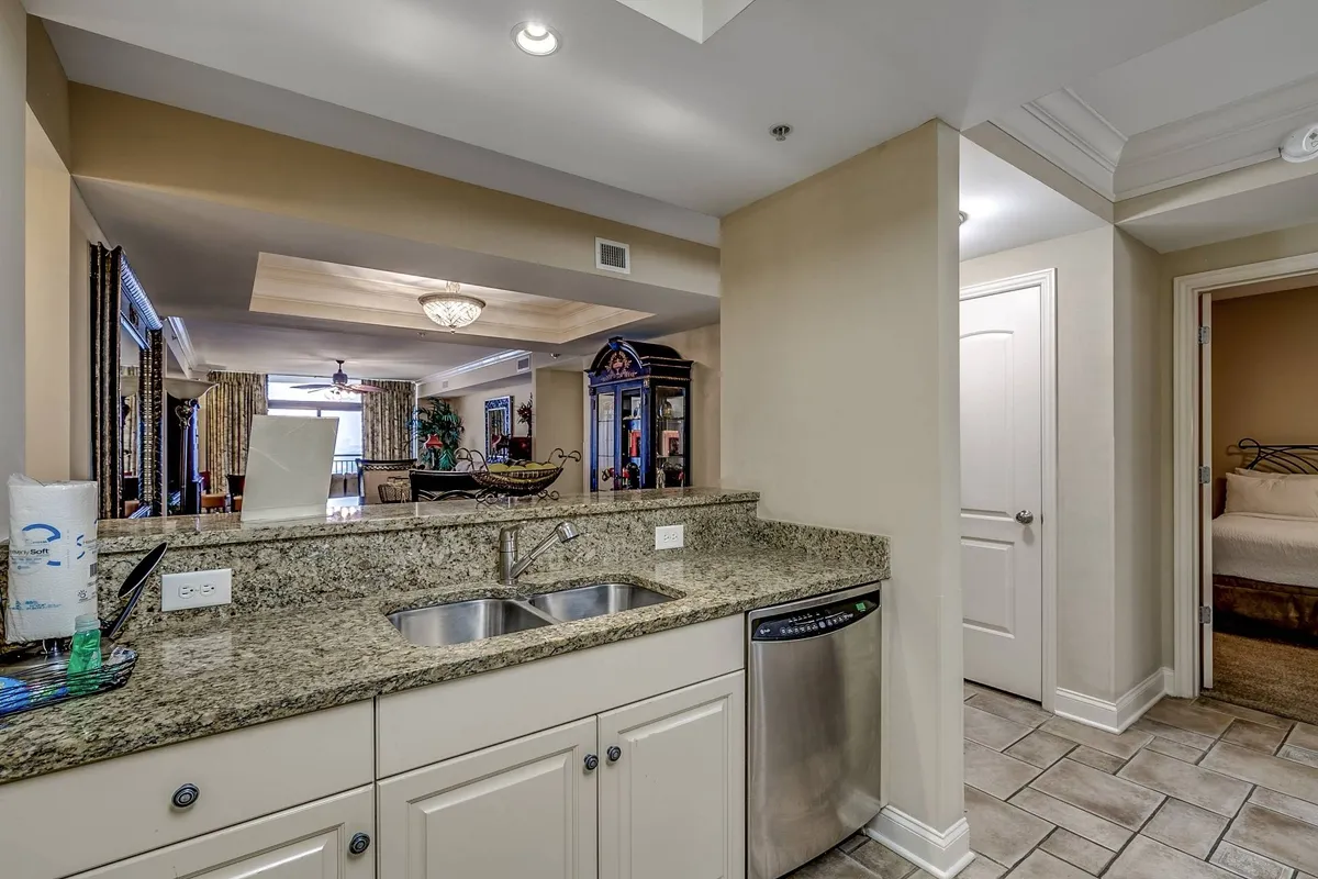 This inviting and practical kitchen scene puts a spotlight on the kitchen sink area, granite countertop, and a visible dishwasher. A mirror reflects the adjoining living space, visually connecting the kitchen to the rest of the home. The layout creates a seamless flow between meal prep and socializing, emphasizing shared meals and the kitchen’s proximity to living and dining areas.