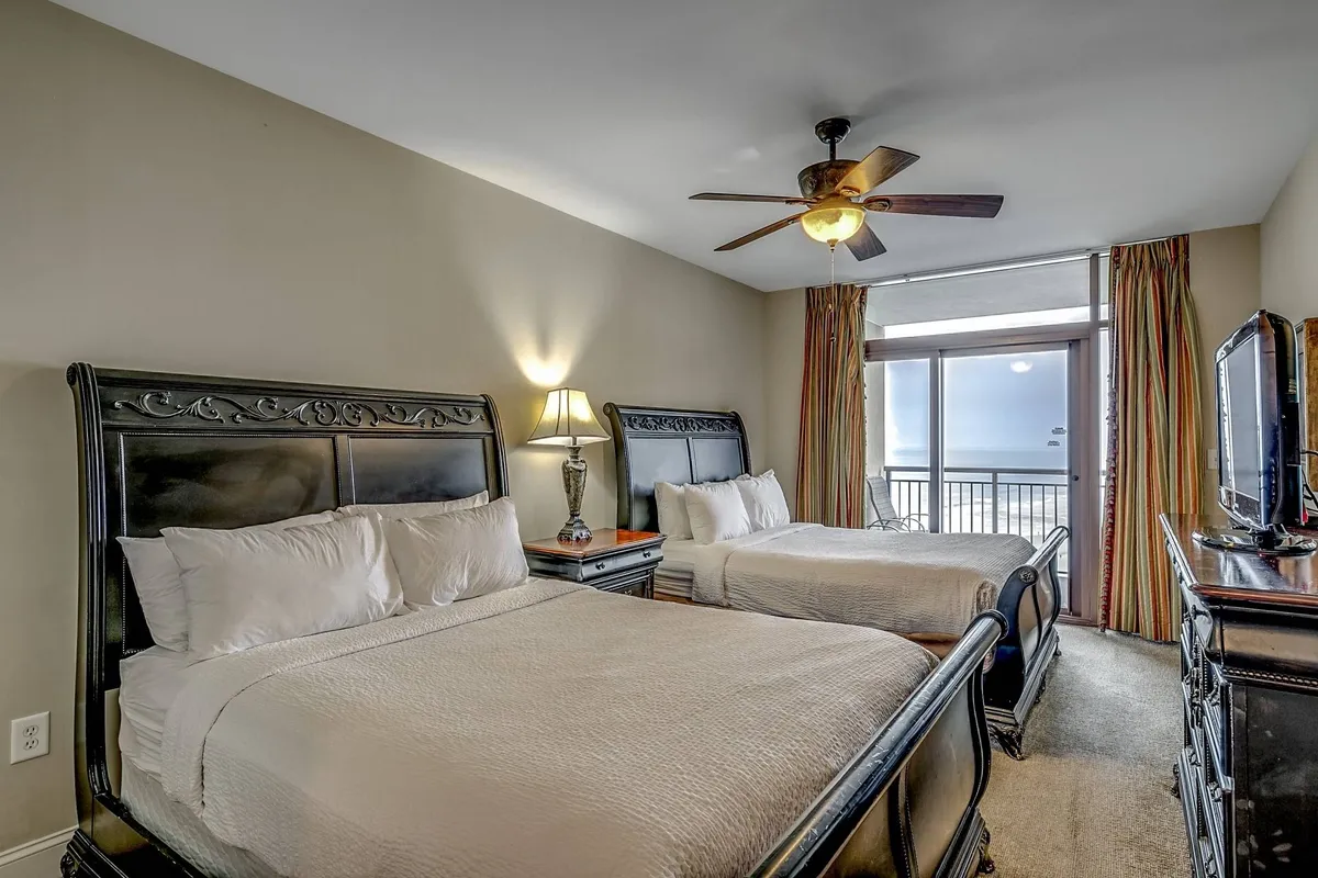 A spacious bedroom with two queen beds, a decorative lamp, curtains, a television, and access to a balcony. The room feels inviting and practical, suitable for families or friends, and highlights options for entertainment and relaxation.