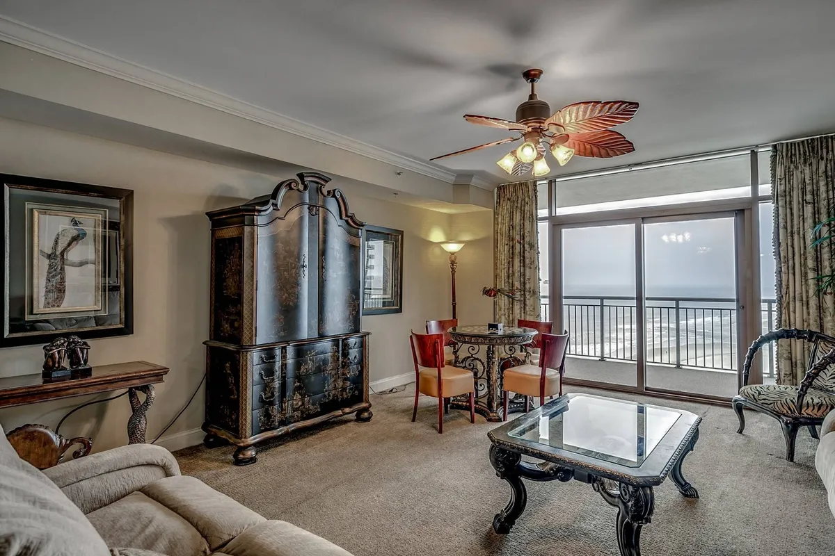 The hero image showcases a stylish dining area featuring an ornate black cabinet, a sleek glass dining table set for gatherings, and a unique leaf-shaped ceiling fan overhead. Sunlight pours in from the balcony, revealing breathtaking ocean views and inviting guests to imagine sunsets enjoyed from the comfort of this elegant space.