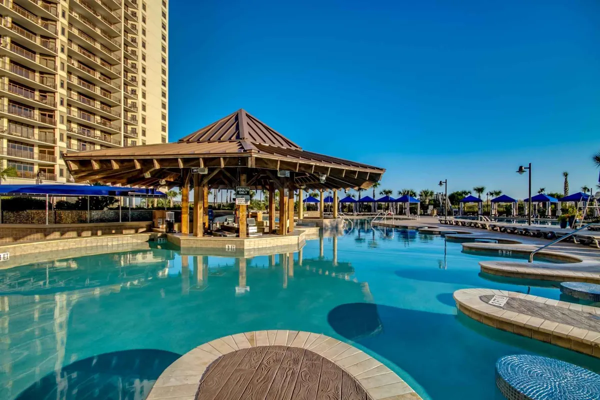 This exterior shot showcases a pool area with a gazebo, plenty of lounge chairs, and sun umbrellas, set against the backdrop of a modern high-rise building. The swim-up bar atmosphere is implied by the gazebo, where guests can socialize and enjoy drinks.